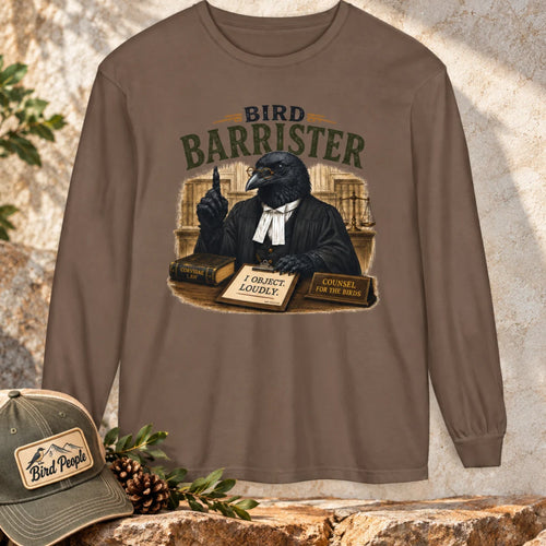 Bird Barrister crow long sleeve t-shirt in Espresso brown, laid flat outdoors with a Bird Peeple trucker hat and pinecones — funny lawyer crow design for birdwatchers.