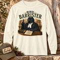 Bird Barrister crow long sleeve t-shirt in Ivory, laid flat outdoors against a rustic wood door with a Bird Peeple trucker hat and pinecones — funny lawyer crow design for birdwatchers.