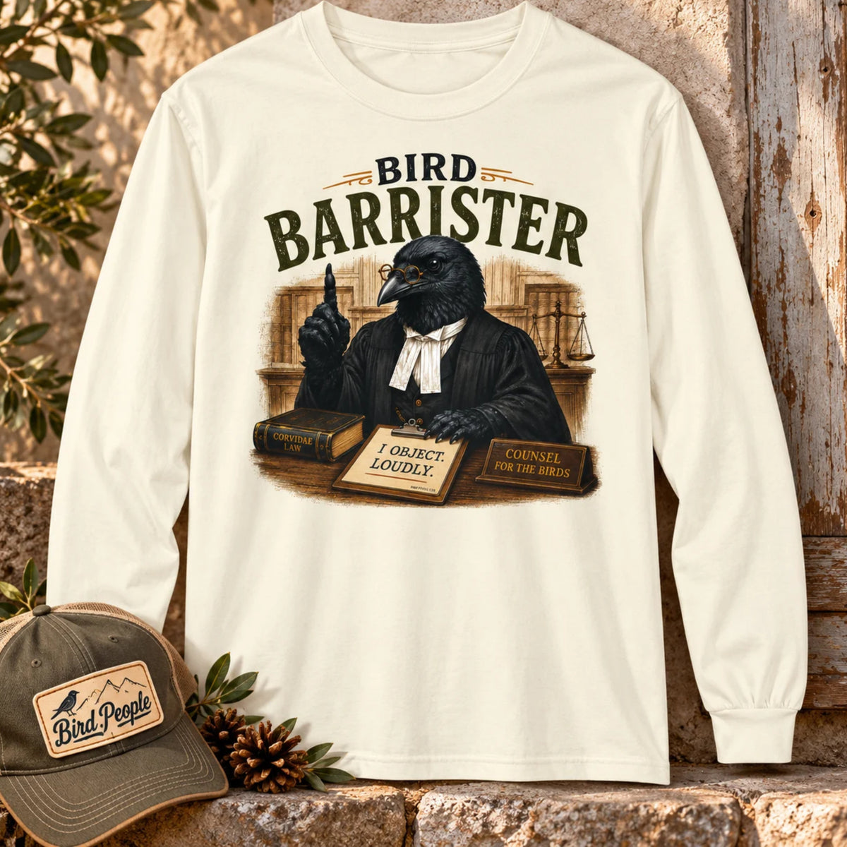 Bird Barrister crow long sleeve t-shirt in Ivory, laid flat outdoors against a rustic wood door with a Bird Peeple trucker hat and pinecones — funny lawyer crow design for birdwatchers.