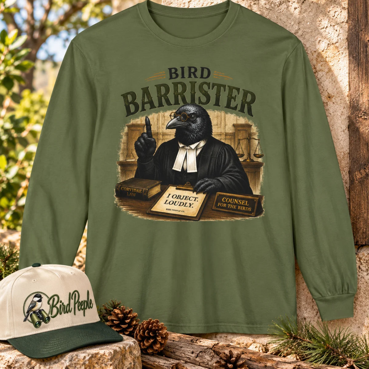 Bird Barrister crow long sleeve t-shirt in Hemp olive green, outdoors with a Bird Peeple chickadee trucker hat and pinecones — funny lawyer crow design for birdwatchers.