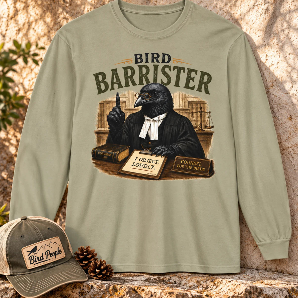 Bird Barrister crow long sleeve t-shirt in Sandstone, laid flat outdoors with a Bird Peeple trucker hat and pinecones — funny lawyer crow design for birdwatchers.