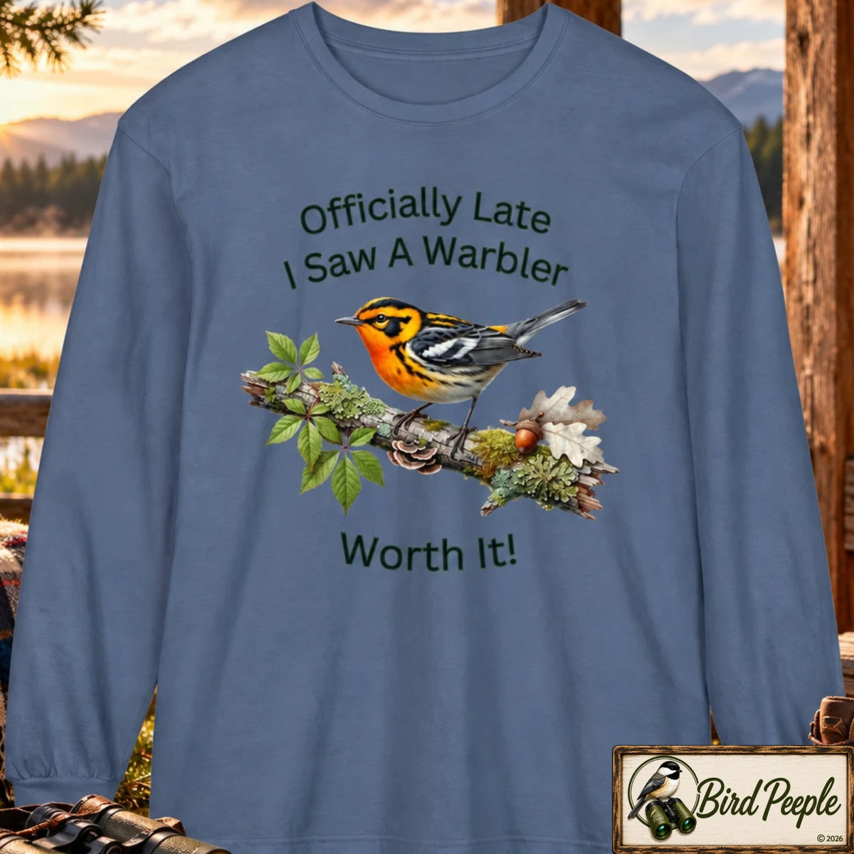 A long-sleeve crewneck t-shirt in blue jean slate blue, featuring a graphic of a warbler on a branch with the text 'Officially Late I Saw A Warbler Worth It!'. The shirt is displayed against a blurred outdoor nature background with a 'Bird Peeple' logo in the bottom right corner. Blackburnian Warbler