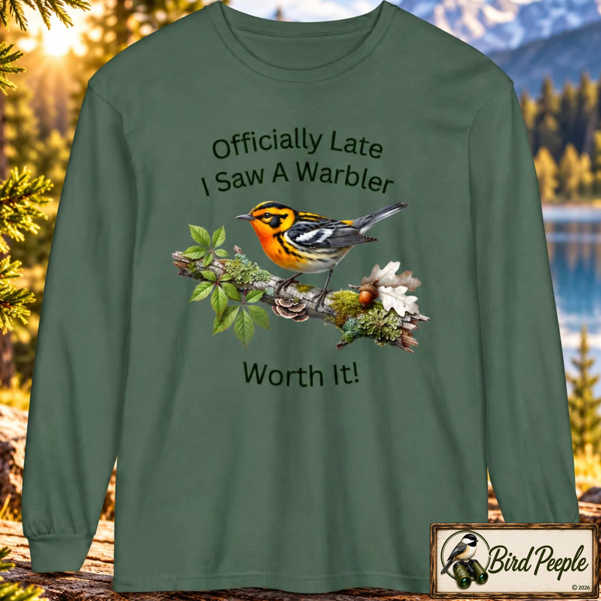 A long-sleeve crewneck t-shirt in Blue Spruce sage green, featuring a graphic of a warbler perched on a branch with the text 'Officially Late I Saw A Warbler Worth It!'. The shirt is displayed against a blurred nature background of a lake and mountains, with a 'Bird Peeple' brand logo in the bottom right corner. Blackburnian Warbler