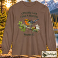 A brown long-sleeve t-shirt with a graphic of a warbler on a branch and the text 'Officially Late I Saw A Warbler Worth It!' displayed against a blurred outdoor nature background. Blackburnian Warbler. A 'Bird Peeple' brand logo is visible in the bottom right corner.