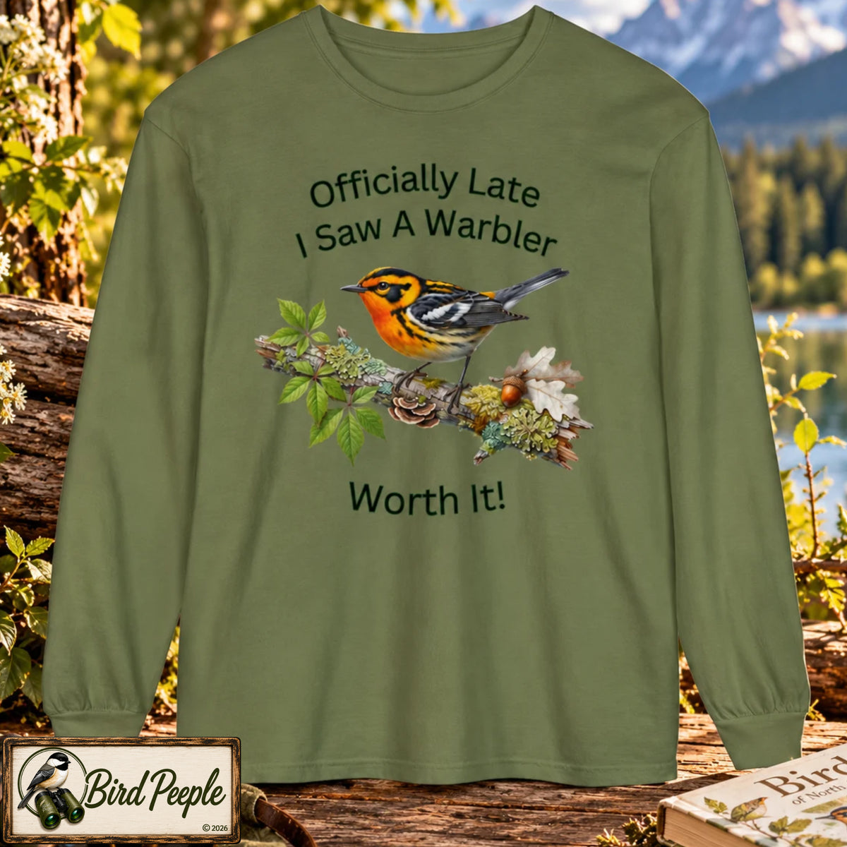 An hemp color olive green long-sleeve t-shirt with a graphic of a warbler on a branch and the text 'Officially Late I Saw A Warbler Worth It!' displayed against a forest and mountain background. Blackburnian Warbler. Bird Peeple logo lower left. 