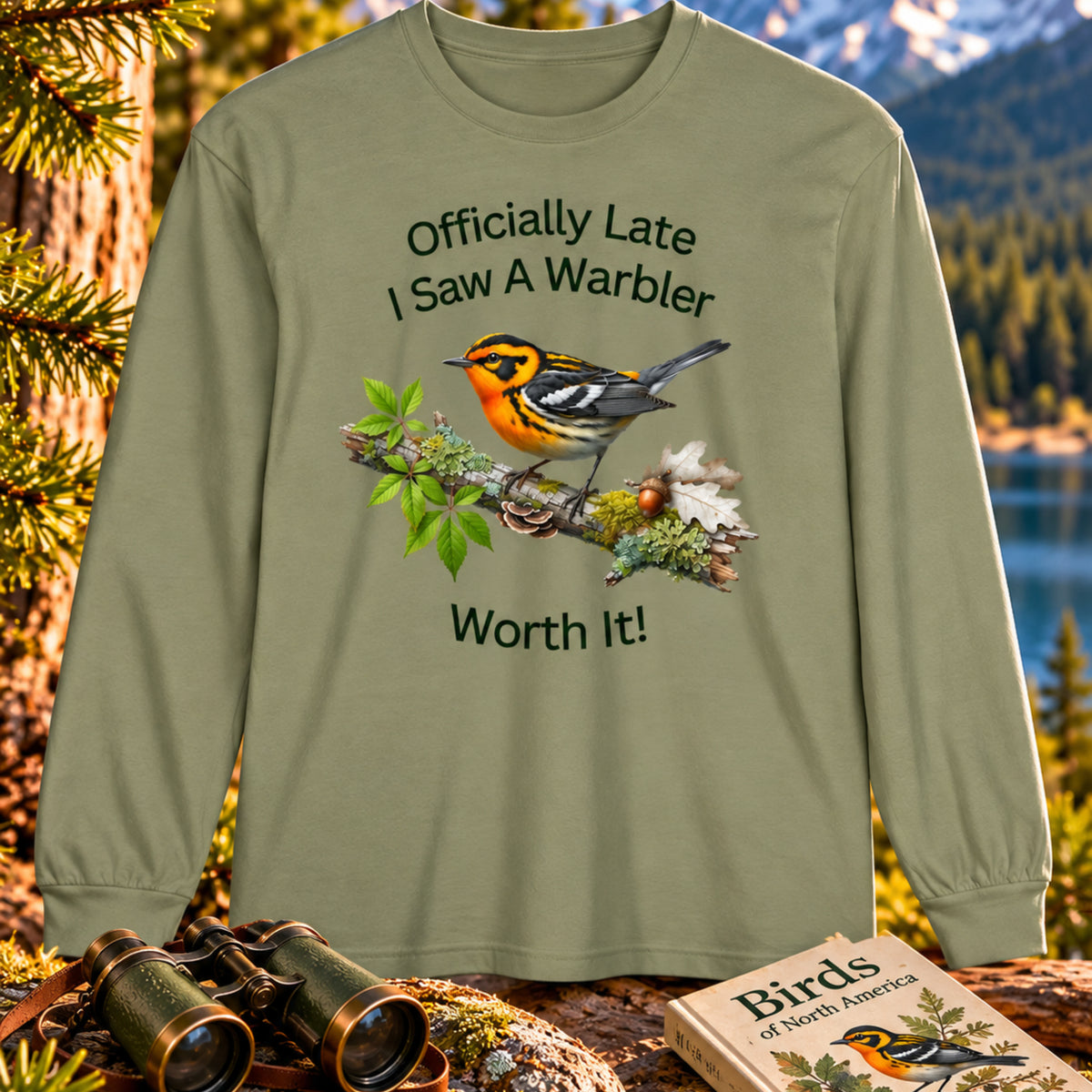 An olive green long-sleeve t-shirt with a graphic of a warbler on a branch and the text 'Officially Late I Saw A Warbler Worth It!' displayed against a scenic outdoor background of a lake and mountains, with binoculars and a book in the foreground. Blackburnian Warbler. Bird Peeple Tee